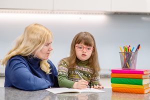 A girl with down syndrome writing in a copybook while woman sitting beside her watches