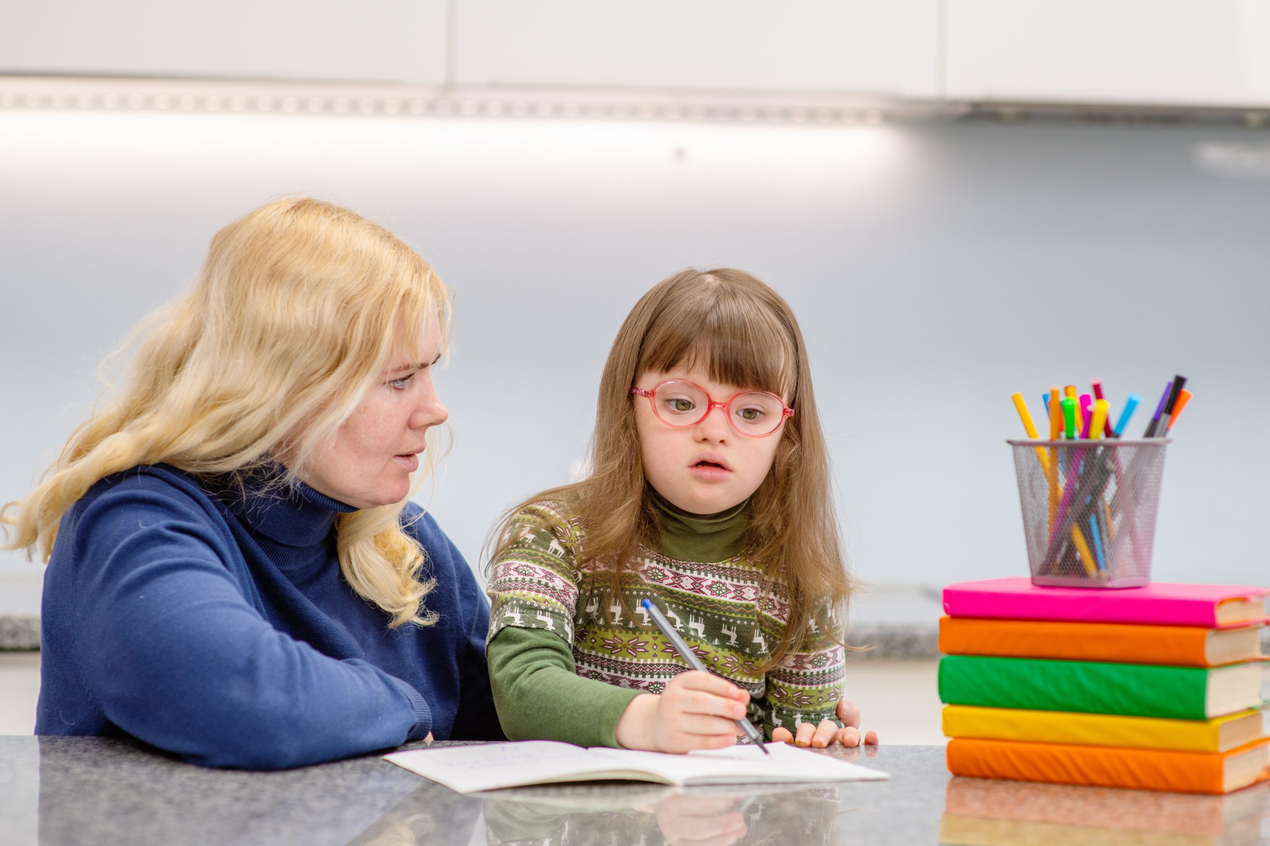 A girl with down syndrome writing in a copybook while woman sitting beside her watches