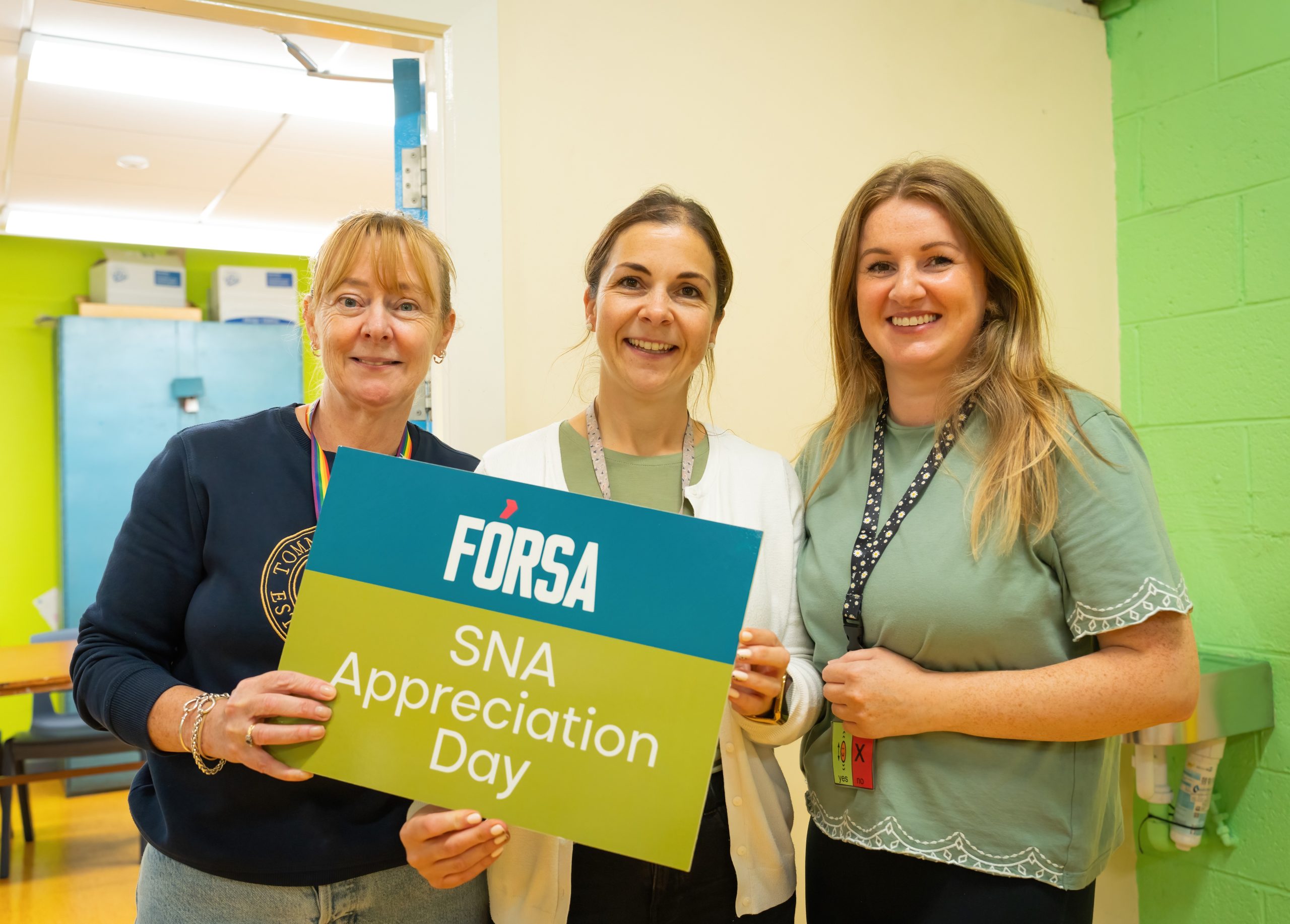 Three SNA members standing in a school hallway holding a sign which reads "Fórsa, SNA appreciation day." The members are all women and the hallway is painted various shades of bright green.