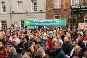 SNA Protest outside Leinster House February 2026. Hundreds in the crowd facing Leinster House. Picture is taken from the Leinster House side of the street facing the protest. At the back of the protest is a long banner that says '#RespectForSNAs' 'United We're Stronger' 'Fórsa SNAs'.
