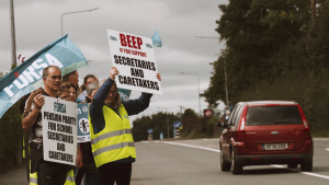 School secretaries and caretakers picketing at on the side of a road.